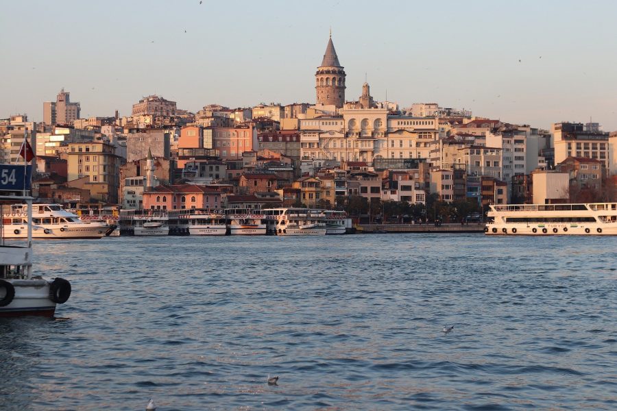 View across calm water of Istanbul cityscape, with Galata Tower standing above the other buildings in the center of the frame. It\'s the golden light hour as sun strikes the buildings.