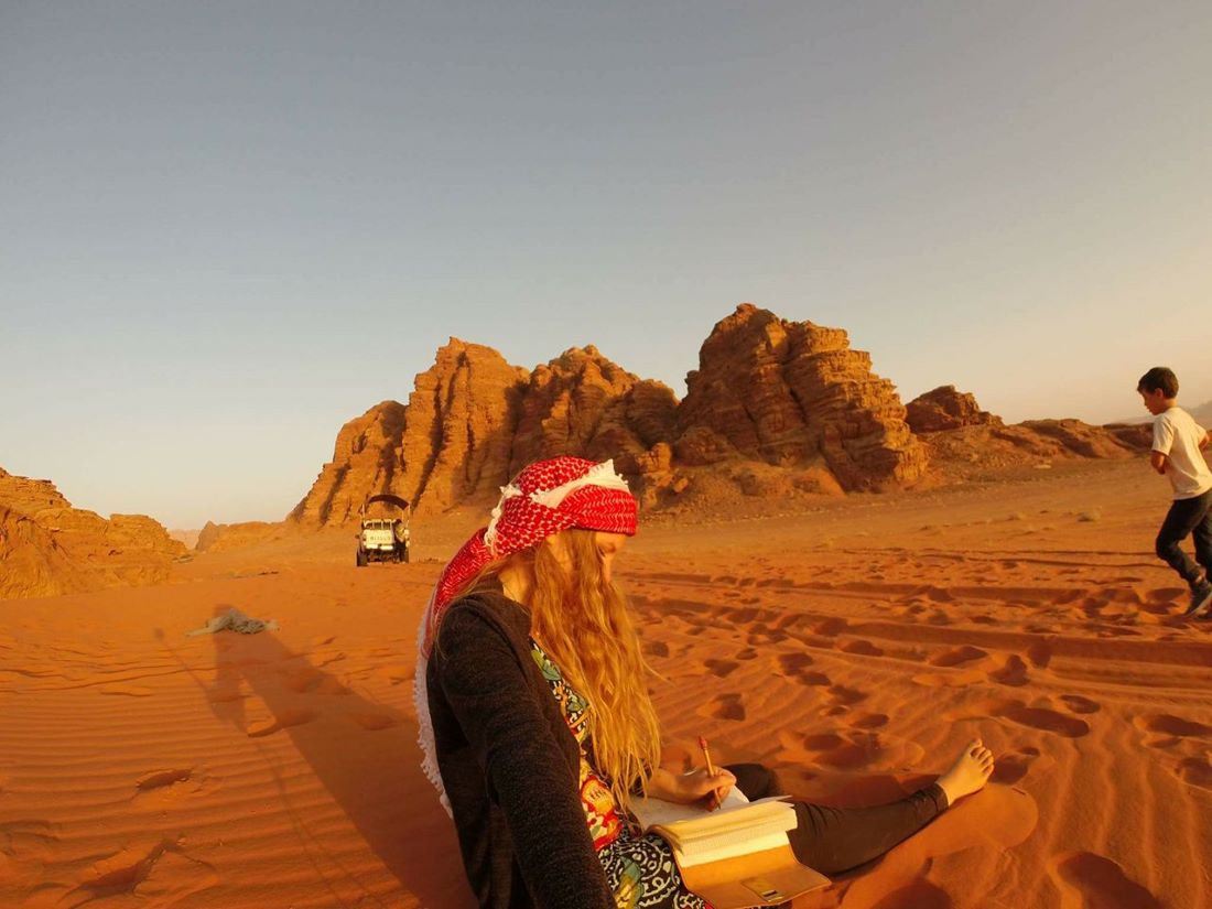 A girl sitting in the Sahara sands writing in her journal while a boy jogs by