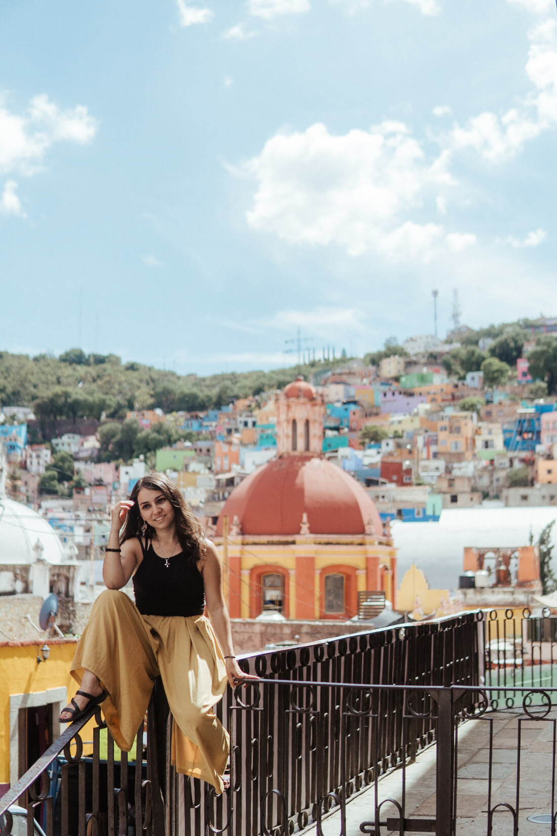 Stacey Valle sitting on a metal railing with a backdrop of colorful houses on the hillside behind her