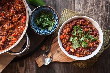 Overhead view of a pot of chili and bowl of chili sitting on separate trivets, with a bowl of chopped green herbs between them.