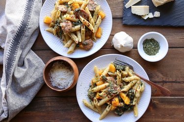 Overhead view of two bowls of penne with sausage and squash, on a wooden table surrounded by a kitchen towel and the dish\'s ingredients.
