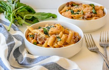 Two white bowls containing pasta, on a white countertop with forks, fresh basil and a kitchen towel.