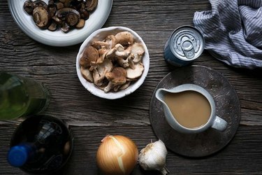 A gravy filled gravy boat next to a bowl of mushrooms, onion, garlic, and a can