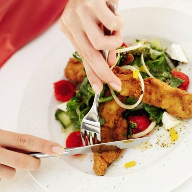 Close-up of a woman\'s hands slicing a piece of fried fish with fork and knife